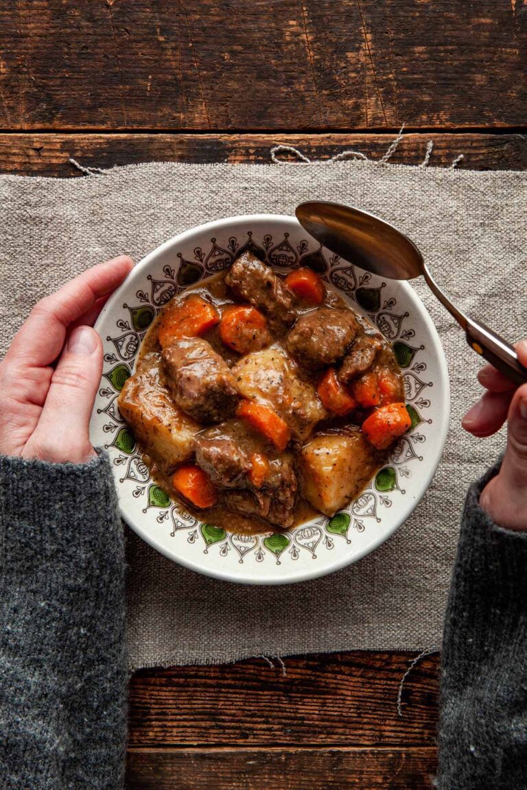 A bowl of Dutch oven beef stew on linen atop a rustic wooden table, with a woman’s hands holding the bowl and a spoon.
