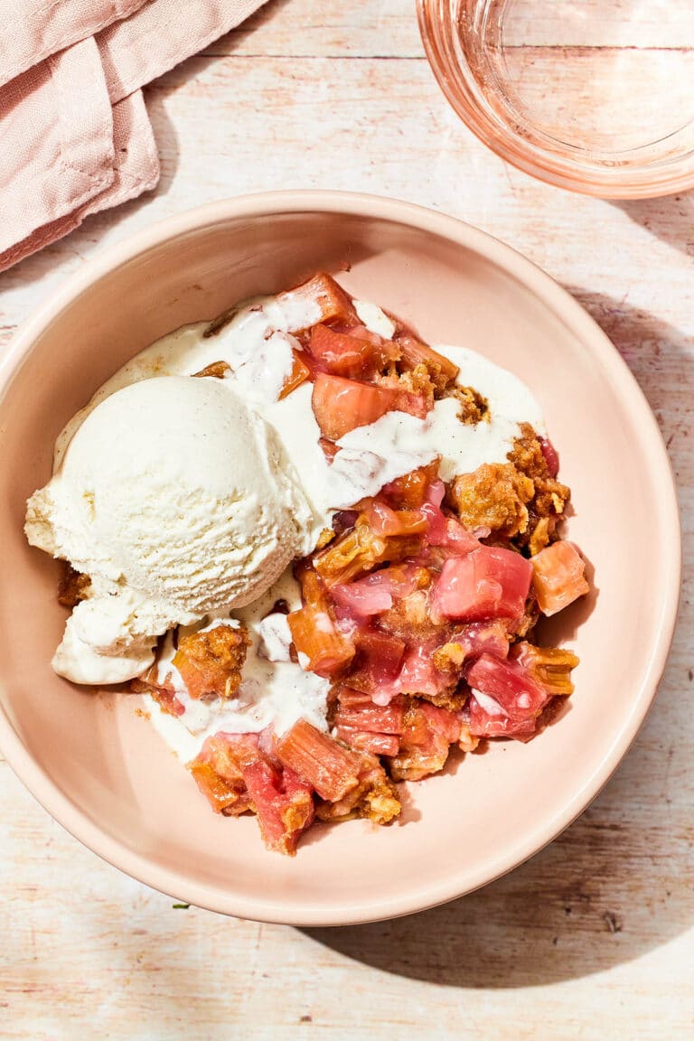 A pink bowl of rhubarb crisp topped with vanilla ice cream on a plank table.
