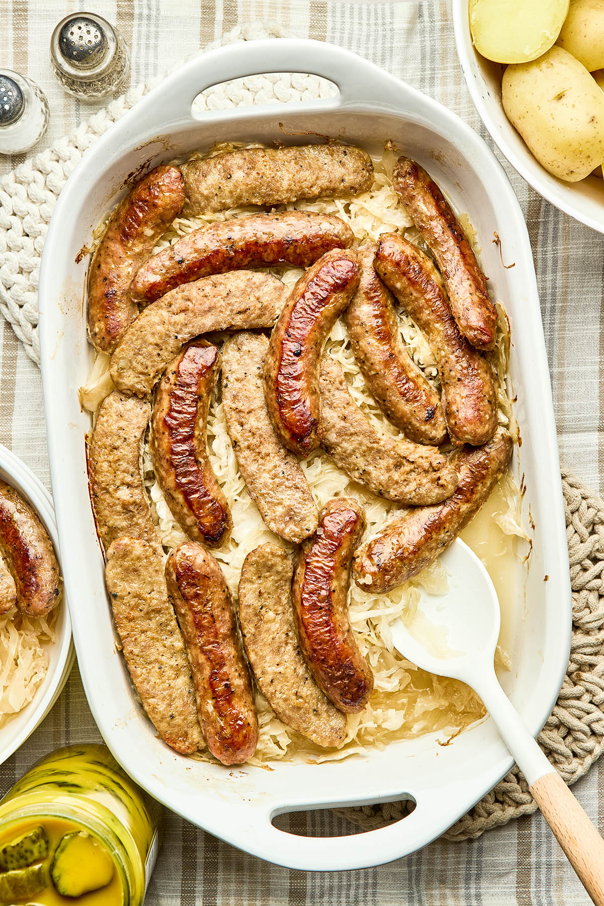 A baking dish of cooked sausage and sauerkraut with a bowl of boiled potatoes, and a jar of mustard pickles nearby.